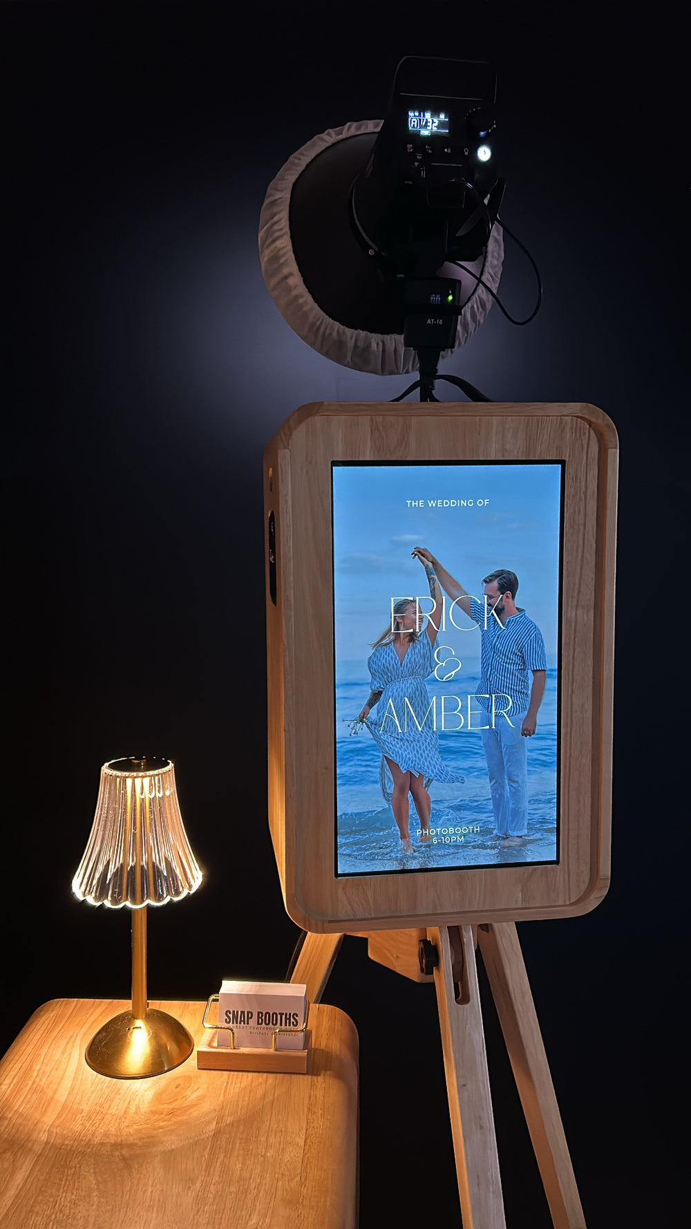 Digital photo booth setup with screen displaying a couple on a beach, surrounded by lighting equipment.