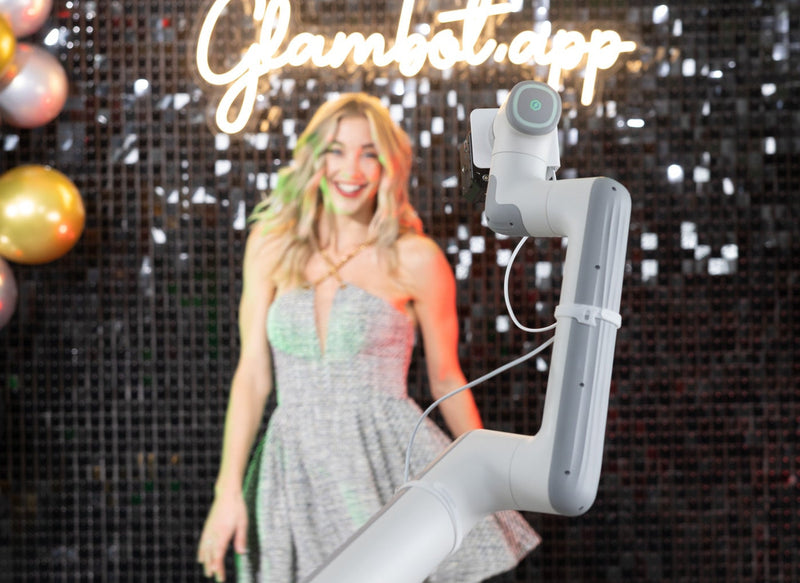 Woman standing next to a robot arm with 'Glambot app' branding in a festive setting.